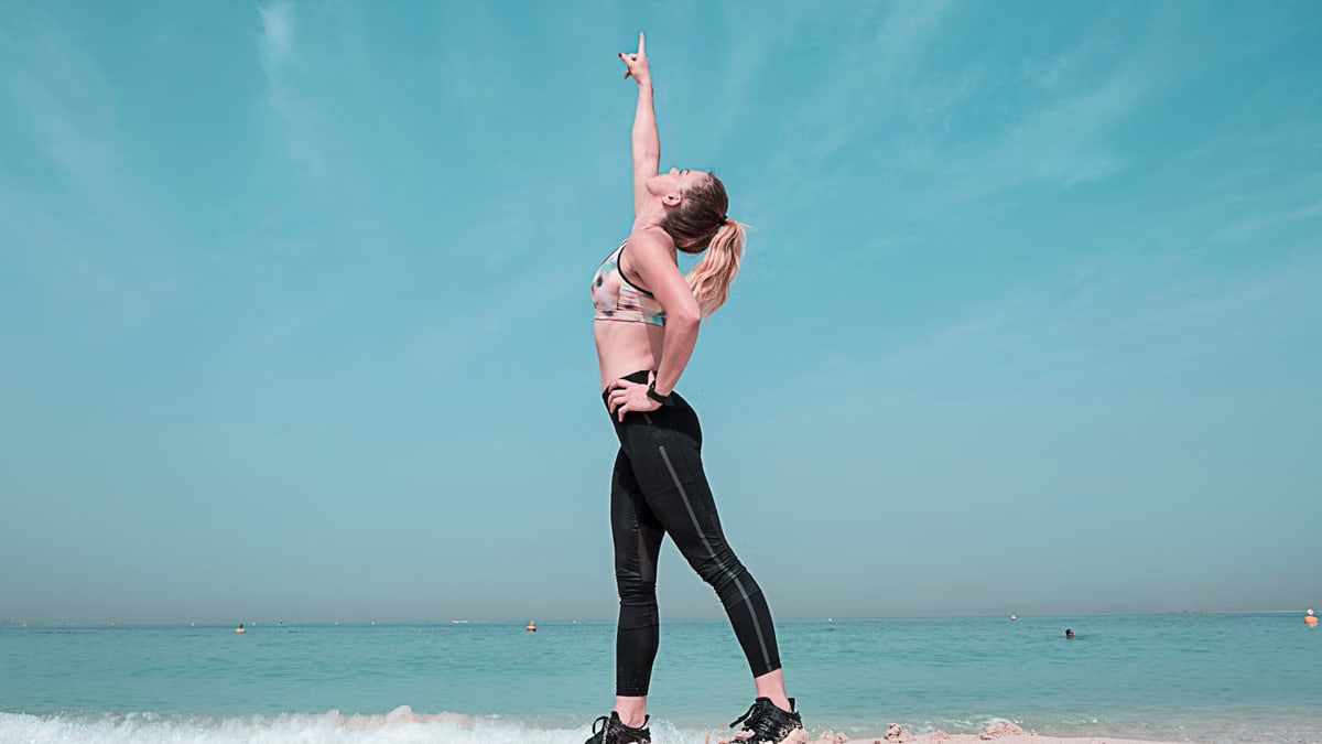 A woman performing a stretching exercise on a sunny beach in Dubai.